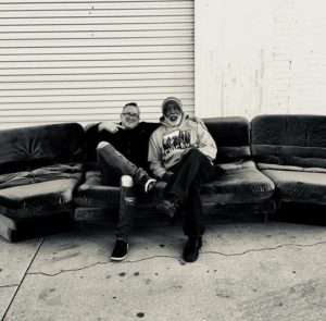 Two professionals from Number2project and Joseph's Upholstery sit side by side on a vintage couch, symbolizing a partnership in furniture restoration." Caption: "Sebastian of Number2project and Joseph of Joseph's Upholstery in Reno, NV, sharing a moment on a mid-century modern Vladimir Kagan couch, showcasing their collaborative resources.